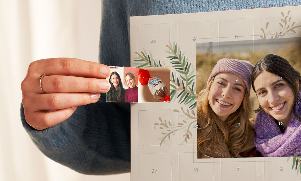 Small hands hold a childhood photo and a chocolate in front of an advent-themed wall picture.