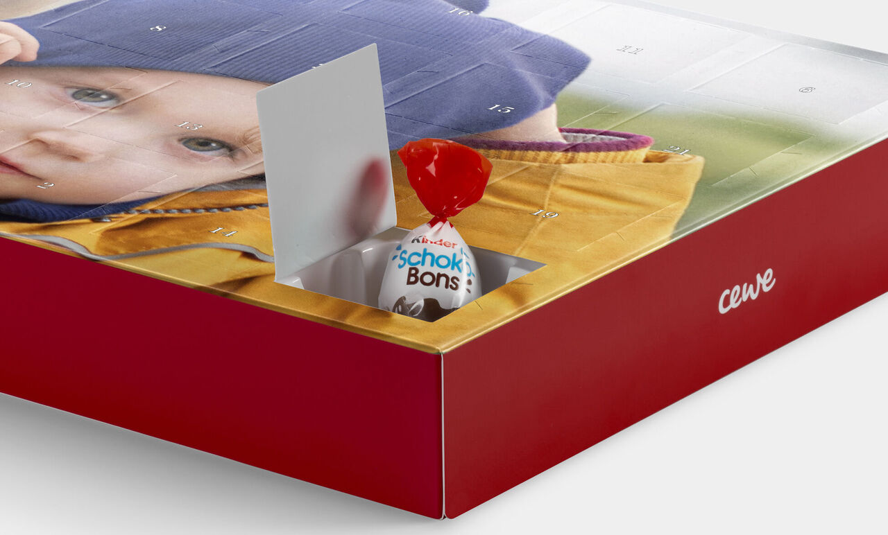 A CEWE photo calendar displays a smiling baby in a yellow jacket. A red chocolate candy is visible.